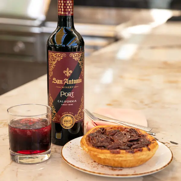 A bottle of San Antonio Winery Port California sweet wine next to a glass of the wine and a pecan tart on a plate, set on a marble countertop with cutlery and a pink napkin in the background.